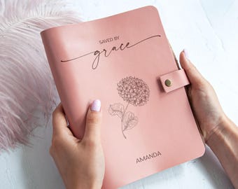 Peut inclure: Un journal en cuir rose poudré doté d'une fermeture à pression, d'une illustration au trait noir représentant une fleur d'hortensia, ainsi que du texte "SAVED BY grace" au-dessus de la fleur et du nom "AMANDA" imprimé en noir en bas.