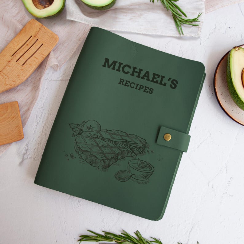 Leaf green leather recipe book with steak illustration and custom engraved name, displayed on a white kitchen background with rosemary and avocado.