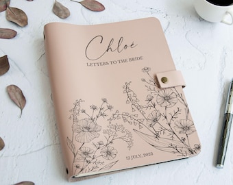 May include: A pale pink journal with the name "Chloe" and the text "Letters to the Bride" printed on the cover. The journal features a floral design and the date "12 July, 2023". A pen and a cup of coffee are next to the journal.