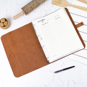 Può includere: Un raccoglitore in pelle marrone contenente una pagina di ricette bianca con intestazioni stampate per ricetta, porzioni, tempo di preparazione, ingredienti, istruzioni e note. Il raccoglitore presenta un meccanismo ad anelli e chiusura.