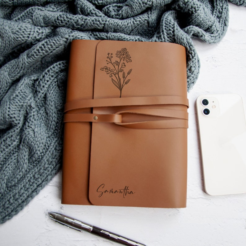 May include: A brown leather journal with a floral design and the name Samantha engraved on the front. The journal has a strap closure and is lying on a white surface.