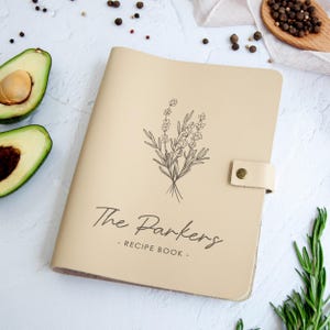 Może przedstawiać: Beżowa książka kucharska z napisem "The Parkers - Recipe Book" i kwiatową ilustracją. Książka ma skórzane zapięcie na pasek z mosiężnym guzikiem. Pokrojone awokado i zioła w tle.