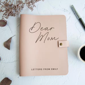 Puede incluir: Un diario de color rosa claro con una cubierta lisa que presenta el texto marrón oscuro Dear Mom en una fuente cursiva y el texto LETTERS FROM EMILY en una fuente sans-serif en la parte inferior. Incluye una correa lateral con un broche.