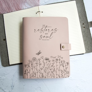 Peut inclure: Un journal rose pâle avec une fermeture à pression, orné du texte "HE restores MY soul PSALM 23:3" et d'une illustration au trait noir représentant un papillon et des fleurs sauvages. Le nom "Maggie Roberts" est inscrit au-dessus du motif floral.