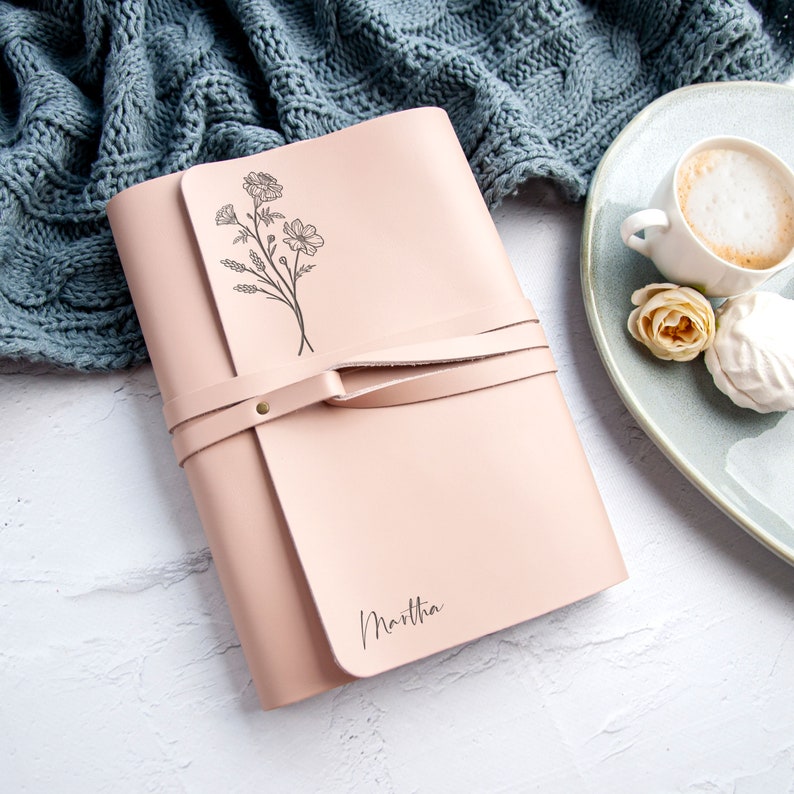 May include: A pink leather journal with a floral design and the name "Marthia" embossed on the cover. The journal has a leather strap closure.
