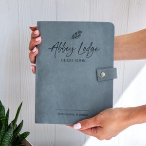 Può includere: Un libro degli ospiti grigio con le parole "Abbey Lodge Guest Book" in corsivo. La copertina presenta un motivo a foglie e una chiusura con cinturino in pelle. La città di Homosassa, Florida, è stampata in basso.