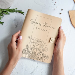 A person's hands holding a beige-colored recipe book with a floral design on the cover.