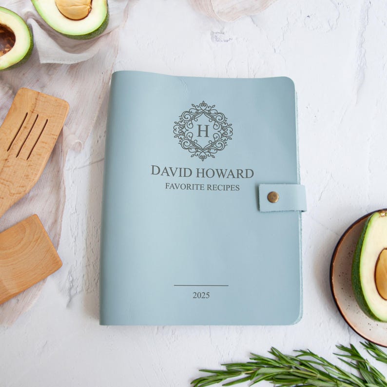 A light blue leather-bound cookbook titled "David Howard Favorite Recipes" with a monogrammed "H" logo, placed on a white surface alongside various kitchen utensils and ingredients.