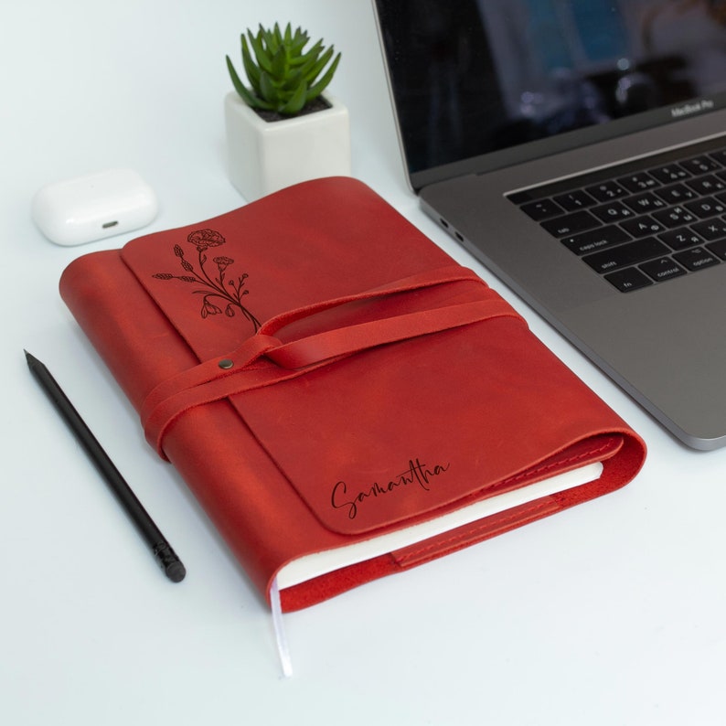 May include: Red leather journal with a floral design and the name Samantha engraved on the front. The journal is closed and has a strap.