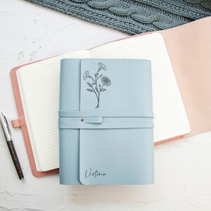 May include: A light blue leather journal with a floral design and the name "Victoria" engraved on the front. The journal has a strap closure and is open to reveal lined pages.