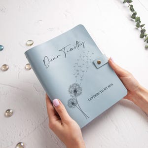 Op de afbeelding: Een lichtblauw notitieboek met de tekst "Dear Timothy" en "Letters to my Son" in zwart schrift. Het notitieboek heeft een paardenbloemillustratie en een drukknoopsluiting.