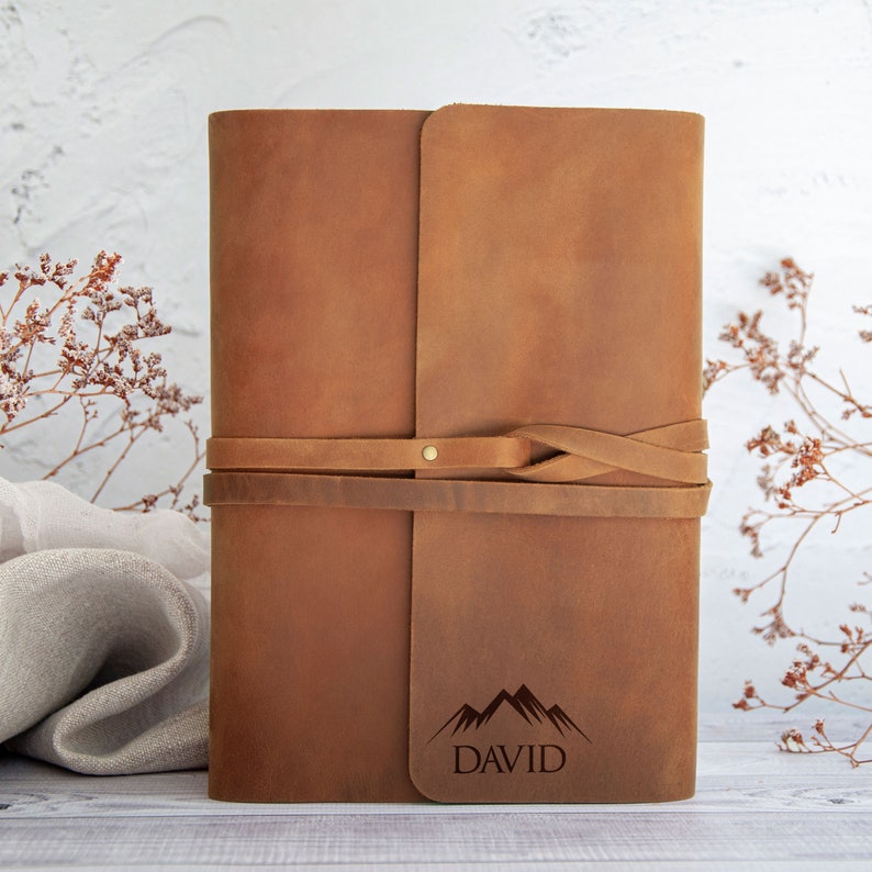 May include: Brown leather journal with a mountain and the name "DAVID" engraved on the front. The journal has a leather strap closure.