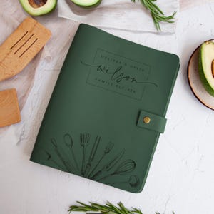 May include: A dark green leather recipe binder featuring a snap closure and a black line art illustration of various kitchen utensils at the bottom. The cover is personalised with the text "MELISSA & DAVID wilson FAMILY RECIPES" in a box.