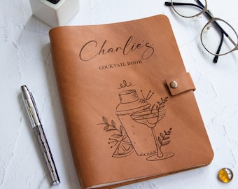 Könnte beinhalten: Ein braunes Cocktailbuch aus Leder mit dem Namen "Charlie's" und den Worten "Cocktail Book" auf dem Einband. Der Einband zeigt auch eine Illustration eines Cocktailshakers und eines Martiniglases. Ein silberner Stift und eine Brille liegen neben dem Buch.