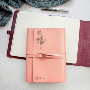 May include: A pink leather journal with a floral design and the name "Brittany" embossed on the front. The journal is closed and has a leather strap.