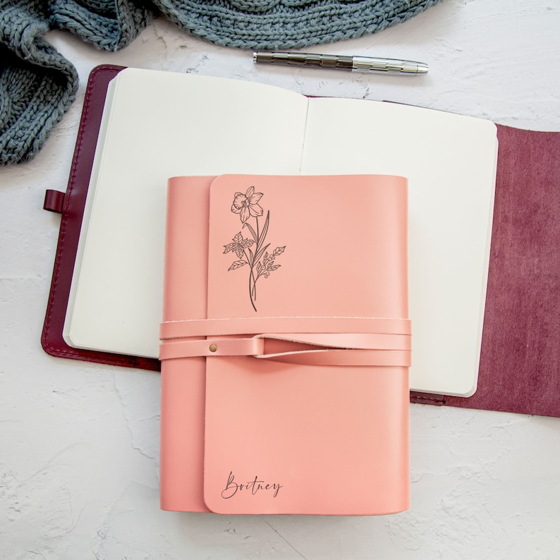 May include: A pink leather journal with a floral design and the name "Brittany" embossed on the front. The journal is closed and has a leather strap.