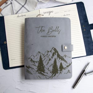 Könnte beinhalten: Ein grauer Rezeptordner mit dem Text "The Bells Family Recipes" und einem Berg- und Baumdesign. Der Ordner ist geöffnet und zeigt linierte Rezeptseiten. Ein Stift liegt daneben.