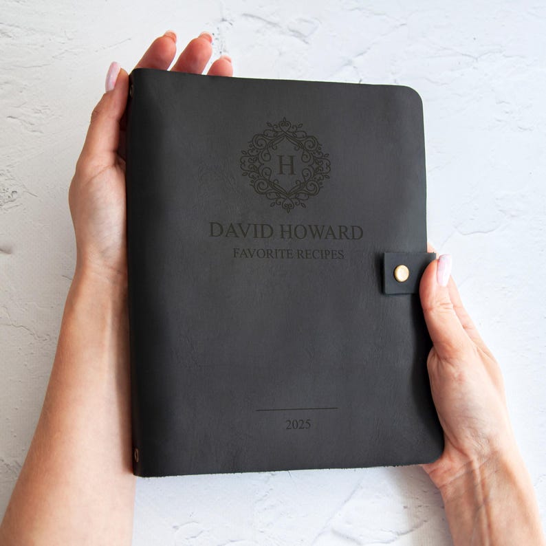 A person's hands holding a black leather-bound notebook with a monogrammed "H" logo and the text "DAVID HOWARD FAVORITE RECIPES" printed on it.