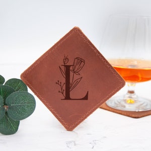 May include: A brown leather coaster with a diamond shape and stitched edges. The coaster features a laser-engraved design of the letter "L" with a floral motif.