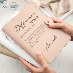 Könnte beinhalten: Ein hellrosa Lederjournal mit den Worten "Difference Maker" und der Definition auf dem Einband. Der Name "Amanda" ist ebenfalls auf dem Einband aufgedruckt. Das Journal hat einen Druckknopfverschluss.