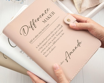 May include: A light pink leather journal with the words "Difference Maker" and the definition printed on the cover. The name "Amanda" is also printed on the cover. The journal has a snap closure and is held open.