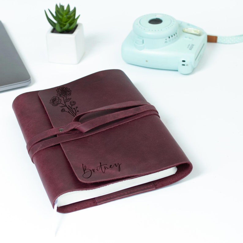 May include: Burgundy leather journal with a floral design and the name "Britney" engraved on the front. The journal has a leather strap closure.
