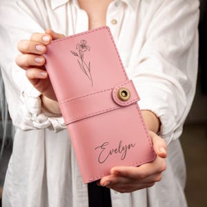 May include: A pink leather wallet with a floral design and the name "Evelyn" in cursive. The wallet has a strap with a brass button closure. The design is a minimalist style, suitable for everyday use.