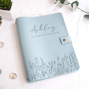 May include: A light blue notebook with a snap closure, featuring the printed text "Ashley" and "LETTERS TO THE BRIDE" in a dark font. The bottom of the cover is decorated with a black line art illustration of various wildflowers and stems.