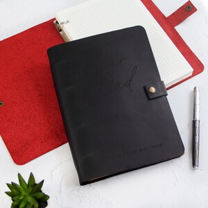May include: A black leather journal cover with a snap closure, embossed with the text "Dear Dad" and "LETTERS FROM MATTHEW". The journal is open to reveal grid-lined paper, and a silver metal pen is positioned to the right of the journal.