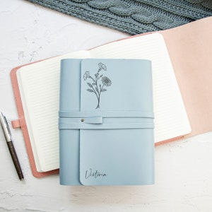 May include: A light blue leather journal with a floral design and the name "Victoria" engraved on the front. The journal has a strap closure and is open to reveal lined pages.