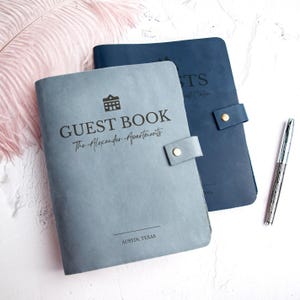 May include: Two leather guest books, one light blue and one navy blue, with silver pen. The light blue book is embossed with "GUEST BOOK" and "The Alexander Apartments" and "AUSTIN, TEXAS". The navy blue book has text on the cover.