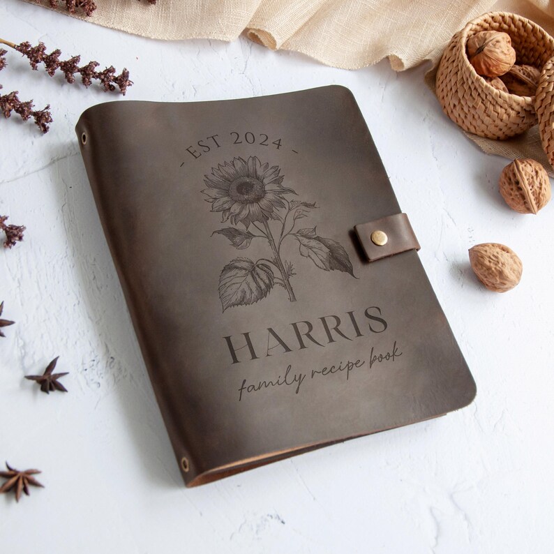 A leather-bound book titled "Harris family recipe book" with a sunflower design, placed on a white surface alongside some nuts and dried flowers.