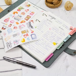 a notebook with handwritten recipes, a pen, and various kitchen-related items such as a bowl, a spoon, and a basket of nuts.