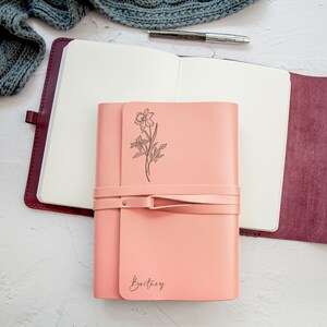 May include: A pink leather journal with a floral design and the name "Brittany" embossed on the front. The journal is closed and has a leather strap.
