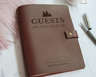 May include: A brown leather guest book with the words "GUESTS of the Evolve Forest Cabin" and "PHOENIX, ARIZONA" printed on the cover. The book has a strap closure with a brass button. A pen and a cup of coffee are next to the book.