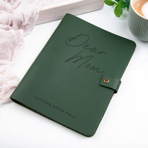 May include: A dark green leather journal featuring a gold-toned snap closure on the side. The cover is embossed with the script text Dear Mom and the block text LETTERS FROM EMILY. The notebook has a smooth, matte finish and rounded corners.