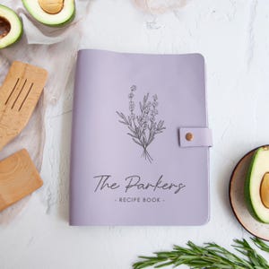 a purple recipe book with a floral design, surrounded by various kitchen utensils and ingredients, such as avocado slices and a wooden spoon.