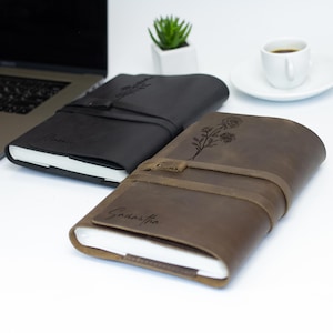 May include: Two personalized leather journals with floral designs and names "Jamie" and "Samantha" engraved on the covers. The journals are closed and lying on a white surface.