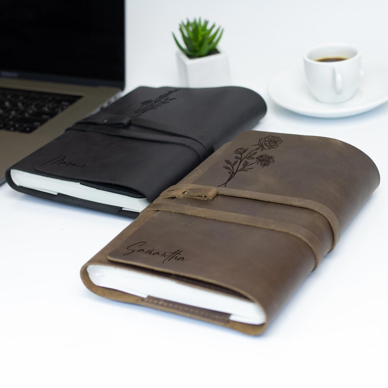 May include: Two personalized leather journals with floral designs and names "Jamie" and "Samantha" engraved on the covers. The journals are closed and lying on a white surface.