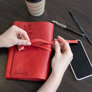 May include: Red leather journal with a floral design and the name "Danielle" engraved on the front. The journal has a leather strap closure and is being tied with a red ribbon.
