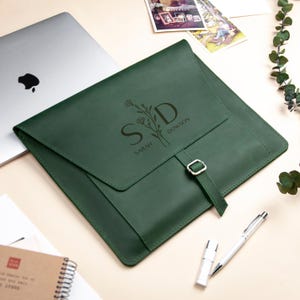 Puede incluir: Una funda para portátil de cuero verde oscuro con cierre de solapa y hebilla. La funda está personalizada con las iniciales "S D" y el nombre "Sarah Dowson" junto con un diseño floral. También se ven un portátil plateado, un bolígrafo y un pequeño cuaderno.