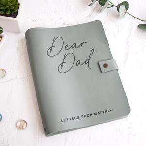 May include: A sage green notebook with a smooth cover featuring the text Dear Dad in a cursive script and the text LETTERS FROM MATTHEW in a sans-serif font. The notebook includes a side strap secured with a small metallic button fastener.