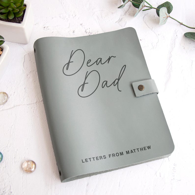 May include: A sage green notebook with a smooth cover featuring the text Dear Dad in a cursive script and the text LETTERS FROM MATTHEW in a sans-serif font. The notebook includes a side strap secured with a small metallic button fastener.