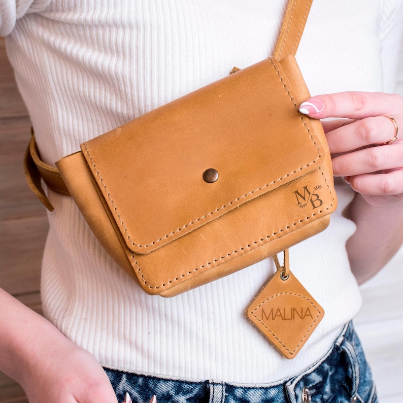 May include: A tan leather fanny pack with a flap closure and a small, diamond-shaped leather tag that says "MALINA". The bag has a metal snap closure and the initials "M&B" are embossed on the front.