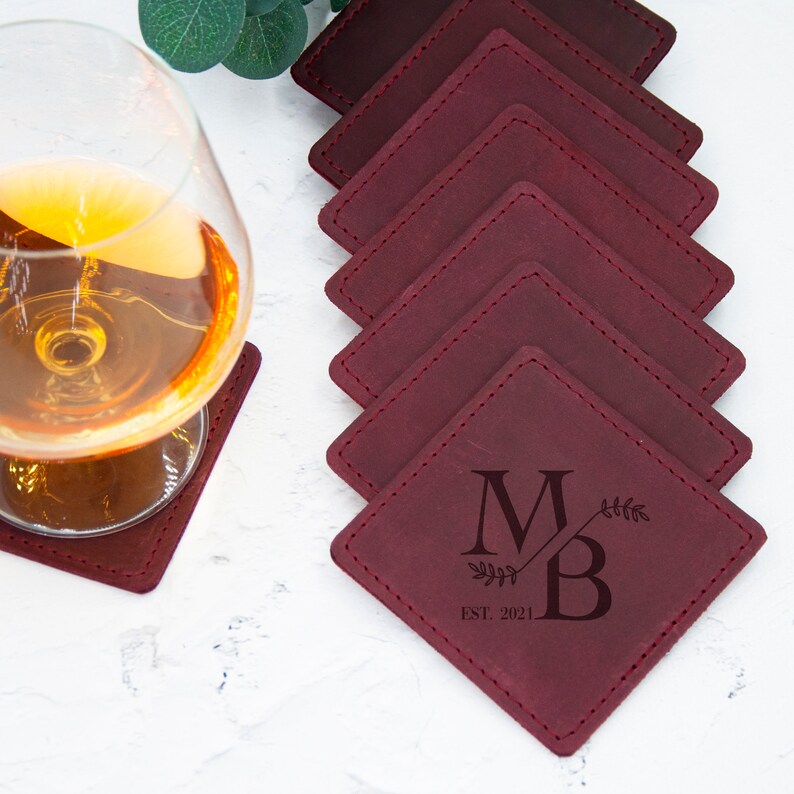 May include: Set of six burgundy leather coasters with stitched edges. The coasters are square and feature a monogram with the initials "MB" and the text "EST. 2021".