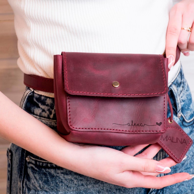 May include: A burgundy leather fanny pack with a flap closure and a small pocket on the front. The pack has the name "alexa" and a heart engraved on the front. A small leather tag with the word "MALINA" is attached to the pack.