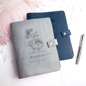 May include: A grey leather journal with a black line art floral design and the name marnie printed on the cover, featuring a side strap with a gold snap button, alongside a dark blue leather journal and a silver metal pen.