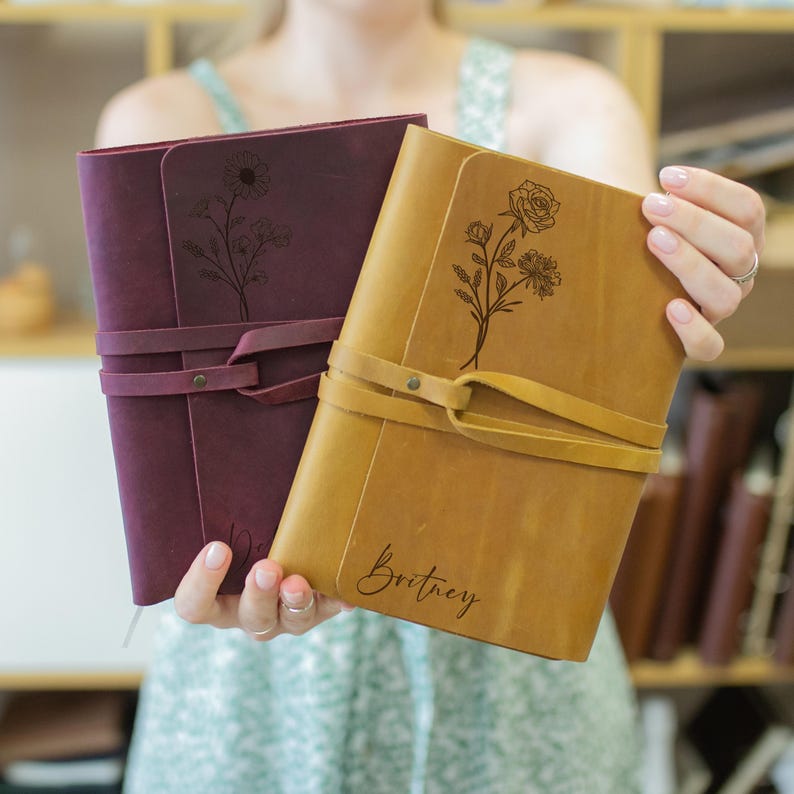 May include: Two leather-bound journals with floral designs and personalized names. The journal on the left is burgundy with a sunflower design and the journal on the right is tan with a rose design. The tan journal is personalized with the name "Britney".