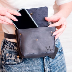 May include: A black leather fanny pack with a phone pocket and a small pocket with the text "M Monica" on it. The fanny pack is worn around the waist of a person wearing blue jeans.