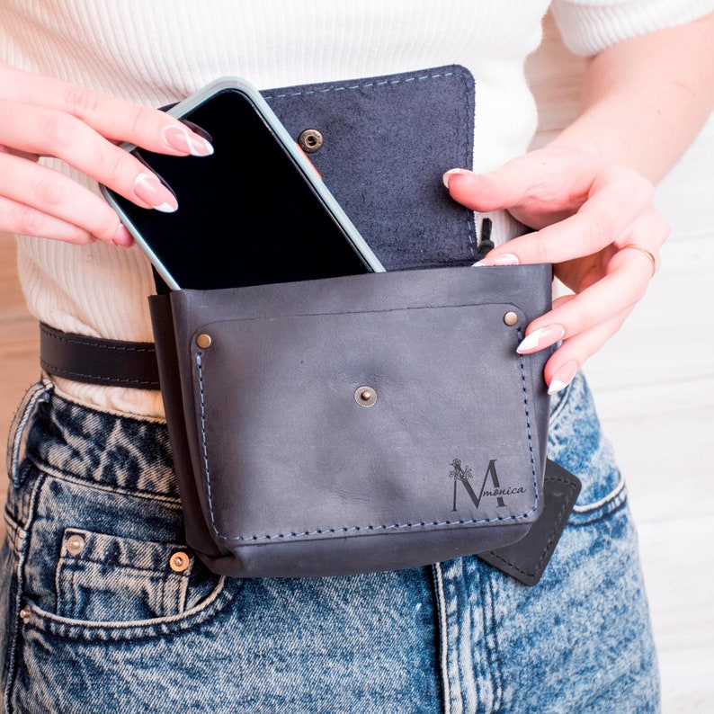May include: A black leather fanny pack with a phone pocket and a small pocket with the text "M Monica" on it. The fanny pack is worn around the waist of a person wearing blue jeans.